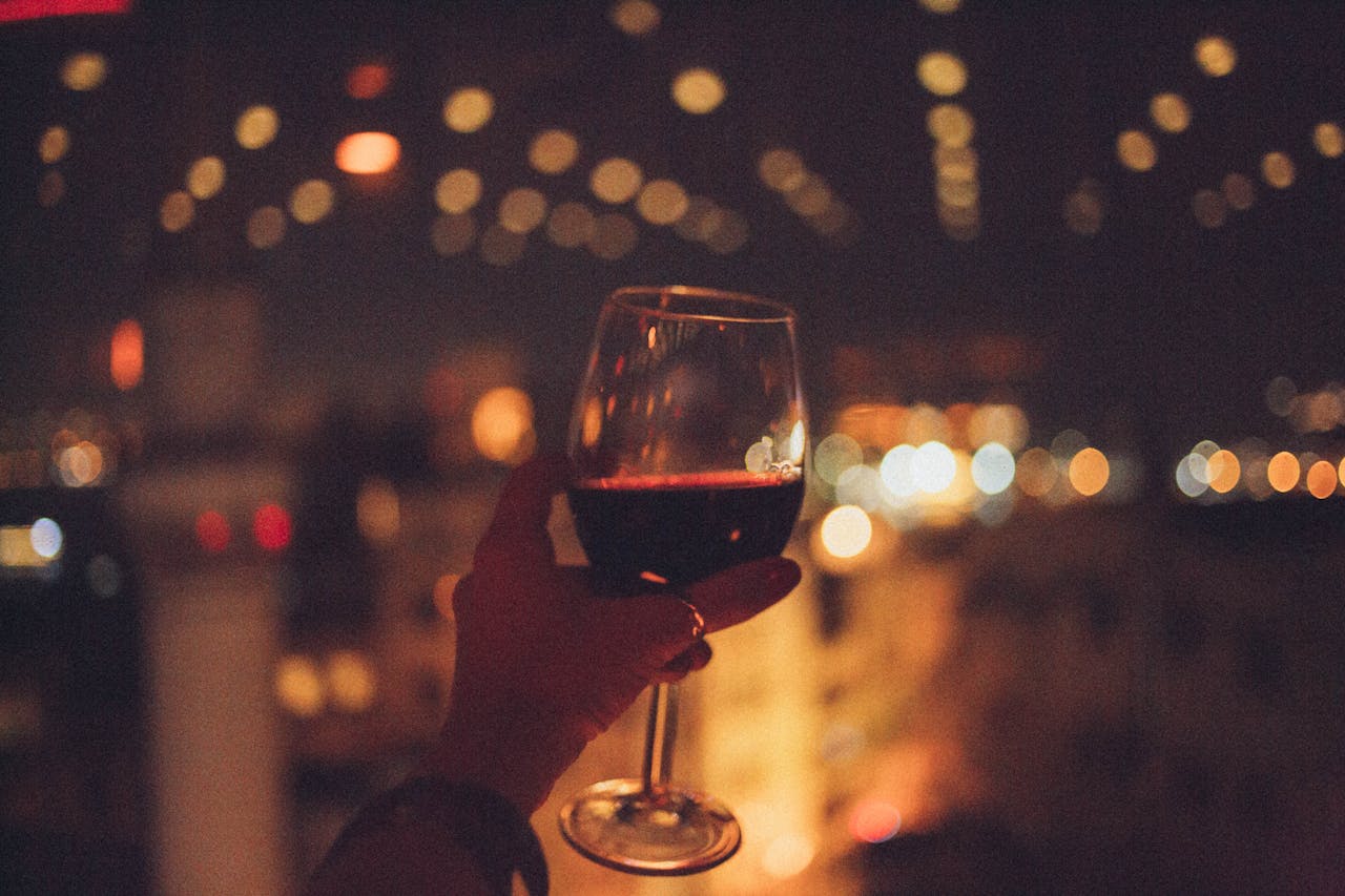 A hand holds a red wine glass with bokeh city lights in a nighttime setting.